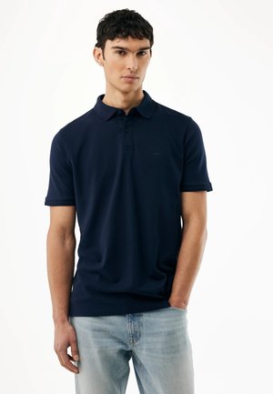 Młody mężczyzna stojący z jedną ręką w kieszeni, ubrany w ciemnogranatową koszulkę polo i jasnoniebieskie dżinsy, na jednolitym białym tle.