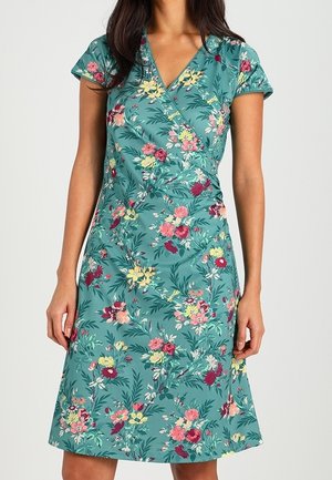Robe portefeuille bleu sarcelle à manches courtes avec des motifs floraux roses, jaunes et rouges, portée par une personne debout, les mains détendues le long du corps.