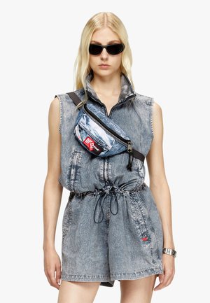 Denim jumpsuit with a standing collar, featuring a cinched waist. Worn with a blue crossbody bag and dark sunglasses. Textured fabric.
