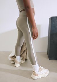 Leggings beiges avec une bande latérale blanche, associés à des chaussettes blanches à côtes et des baskets de couleur claire. Texture lisse et design ajusté.