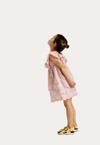 Robe rose clair avec des motifs floraux brodés, ourlet festonné et manches à volants. Sandales jaunes avec des accents noirs et des sangles à boucle.