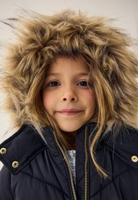 Doudoune noire rembourrée avec une capuche doublée en fausse fourrure, des fermetures éclair dorées et des détails de couture marquants. Texture lisse et coupe ajustée.