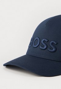 Marineblå baseballcap laget av stoff med en brodert "BOSS"-logo på fronten. Har en buet skjerm og teksturert overflate.