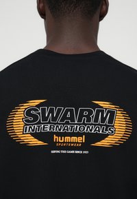 Tricou negru din bumbac cu un grafic mare alb și portocaliu pe spate, având textul "SWARM INTERNATIONALS" și "hummel sportswear."