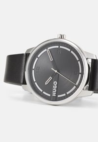 Montre ronde avec un bracelet en cuir noir, quincaillerie en argent, et un cadran texturé gris foncé comportant des accents blancs et le nom de la marque "HUGO."
