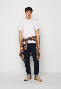 Camiseta blanca de cuello redondo, jeans azul oscuro, camisa de cuadros marrón anudada a la cintura, botines beige con paneles elásticos en los lados.