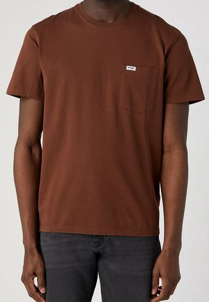 Man draagt een effen bruine korte mouw t-shirt met een borstzakje met het label Wrangler, gecombineerd met donkergrijze broek.