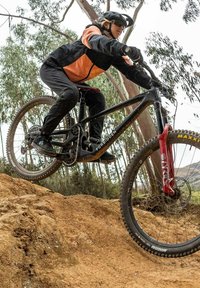 Horský bicykl s černým rámem a červenou přední vidlicí, vybavený širokými pneumatikami a řetězem. Jezdec v oranžové a černé bundě.