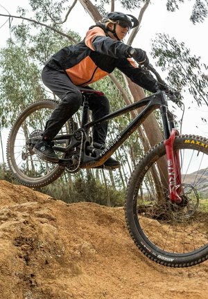 Horský bicykl s černým rámem a červenou přední vidlicí, vybavený širokými pneumatikami a řetězem. Jezdec v oranžové a černé bundě.