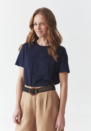 T-shirt blu navy a maniche corte con un design di foglia ricamata e testurizzata. Abbinato a pantaloni beige a vita alta e una cintura con motivo.