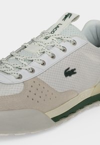 Λευκό και μπεζ αθλητικό παπούτσι Lacoste με πάνελ από δίχτυ και σουέτ, λευκά κορδόνια, πράσινο λογότυπο κροκόδειλου και πράσινες και κρεμ λεπτομέρειες στη σόλα.