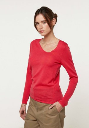 Vrouw met donker haar in een lage knot, gekleed in een rode top met lange mouwen en V-hals en beige broek, staand met één hand in de broekzak tegen een witte achtergrond.