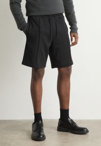 Shorts de sport noirs avec une taille élastique et un cordon de serrage. Portés avec des chaussures habillées noires et des chaussettes, complétés par un pull sombre.