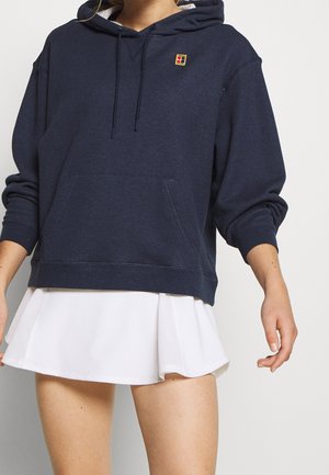 Personne portant un sweat à capuche bleu foncé avec une poche frontale et un petit logo, associé à une jupe de tennis plissée blanche sur un fond uni.