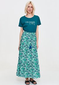 Turkusowa koszulka z białym napisem, zestawiona z długą, wzorzystą spódnicą w odcieniach zieleni i niebieskiego, z troczkiem i detalem w postaci frędzla. Czarne sandały.