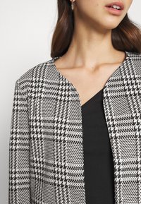 Chaqueta con patrón de pata de gallo en blanco y negro, con un escote redondeado y tejido texturizado, combinada con una camiseta negra debajo.