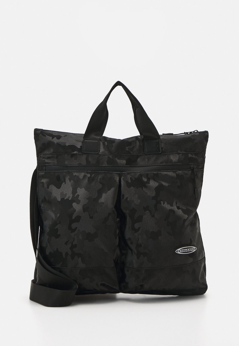Sac cabas noir à motif camouflage avec doubles poignées supérieures, poche zippée avant et bandoulière réglable, avec un petit écusson logo.