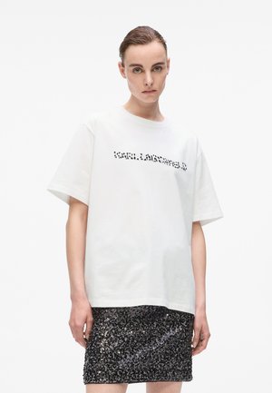 Una giovane donna con i capelli slicked-back indossa una maglietta bianca oversize e una mini gonna nera con texture, in piedi davanti a uno sfondo bianco.
