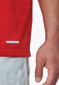 Camiseta de algodón roja con cuello redondo, manga corta y una etiqueta de logo plateada en el dobladillo, combinada con pantalones cortos de mezclilla claros.