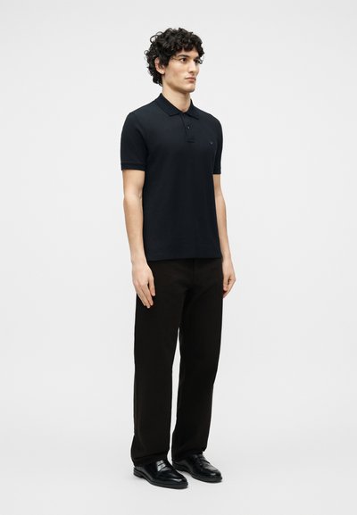 Νεαρός άνδρας με σγουρά μαλλιά όρθιος, φορώντας μαύρο πόλο με κοντά μανίκια, μαύρο παντελόνι και μαύρα επίσημα παπούτσια σε απλό λευκό φόντο.