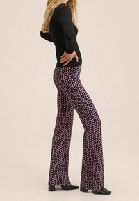 Kobieta w czarnym prążkowanym swetrze i purpurowych spodniach z wysokim stanem w czarno-różowy kwiatowy wzór, stojąca w profilu z rękami na biodrach.