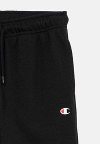 Schwarze Jogginghose mit elastischem Bund, Seitentaschen und einem kleinen weißen und roten Logo am linken Bein. Weiches Baumwollmischgewebe.