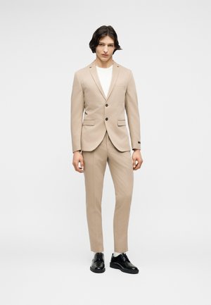 Νεαρός άνδρας που στέκεται, φορώντας μπεζ κοστούμι με σακάκι δύο κουμπιών, λευκό πουκάμισο, λευκές κάλτσες και μαύρα επίσημα παπούτσια, σε λευκό φόντο.