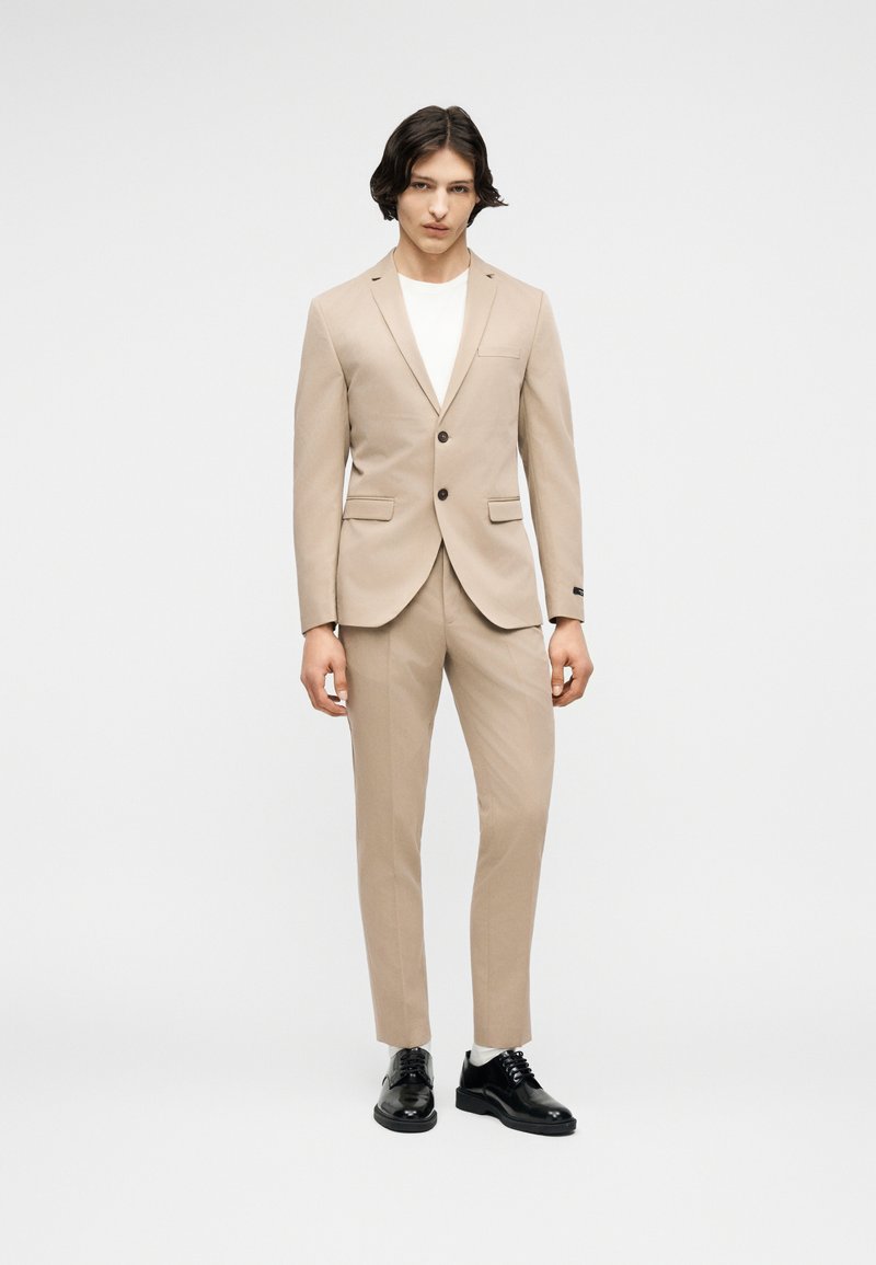 Νεαρός άνδρας που στέκεται, φορώντας μπεζ κοστούμι με σακάκι δύο κουμπιών, λευκό πουκάμισο, λευκές κάλτσες και μαύρα επίσημα παπούτσια, σε λευκό φόντο.