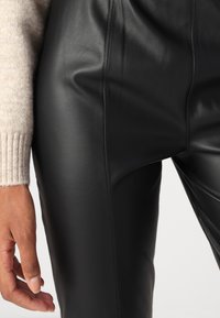 Zwarte leren broek met een gladde textuur, voorzien van naaddetails. Een lichtgekleurde gebreide trui is gedeeltelijk zichtbaar aan de bovenkant.