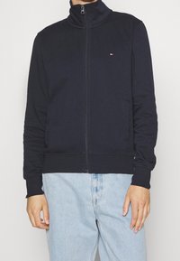 Uomo che indossa una giacca zip-up blu navy con un piccolo logo sul petto e jeans azzurri chiari su uno sfondo semplice.