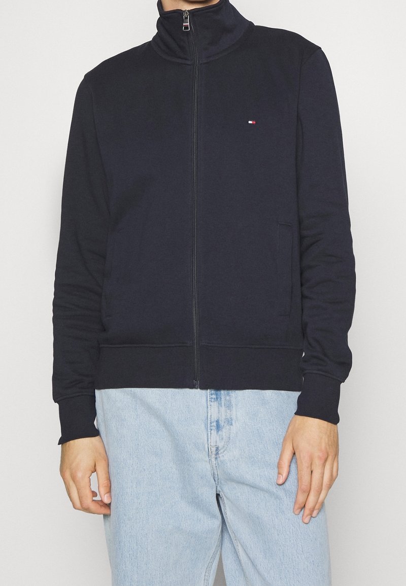 Uomo che indossa una giacca zip-up blu navy con un piccolo logo sul petto e jeans azzurri chiari su uno sfondo semplice.