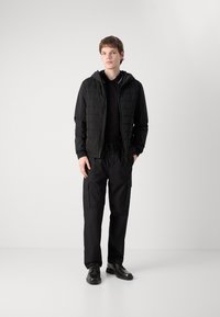 Giacca nera trapuntata con cappuccio sopra una polo nera, abbinata a pantaloni cargo neri larghi e scarpe nere stringate. Tessitura liscia in tutto.