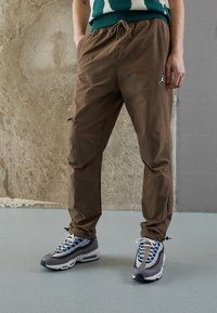 Bruine, lichtgewicht joggingbroeken met elastische tailleband en trekkoord; voorzien van een logo-accent op het been. Gecombineerd met grijze en blauwe sportschoenen.