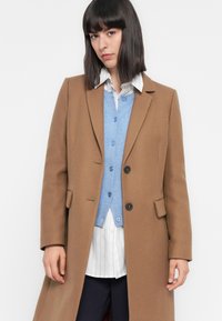 Femme avec des cheveux foncés et raides portant un manteau en laine beige par-dessus un cardigan bleu et une chemise blanche à rayures, sur un fond blanc uni.