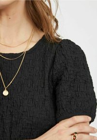 Top nero texturizzato con scollatura rotonda, maniche corte a sbuffo, collane d'oro sovrapposte e un anello decorativo sulla mano.
