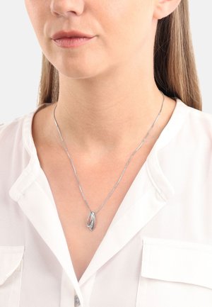 Collana d'argento con un pendente caratterizzato da un design di foglie intrecciate, indossata sopra una camicia bianca. La catena è delicata e slanciata.