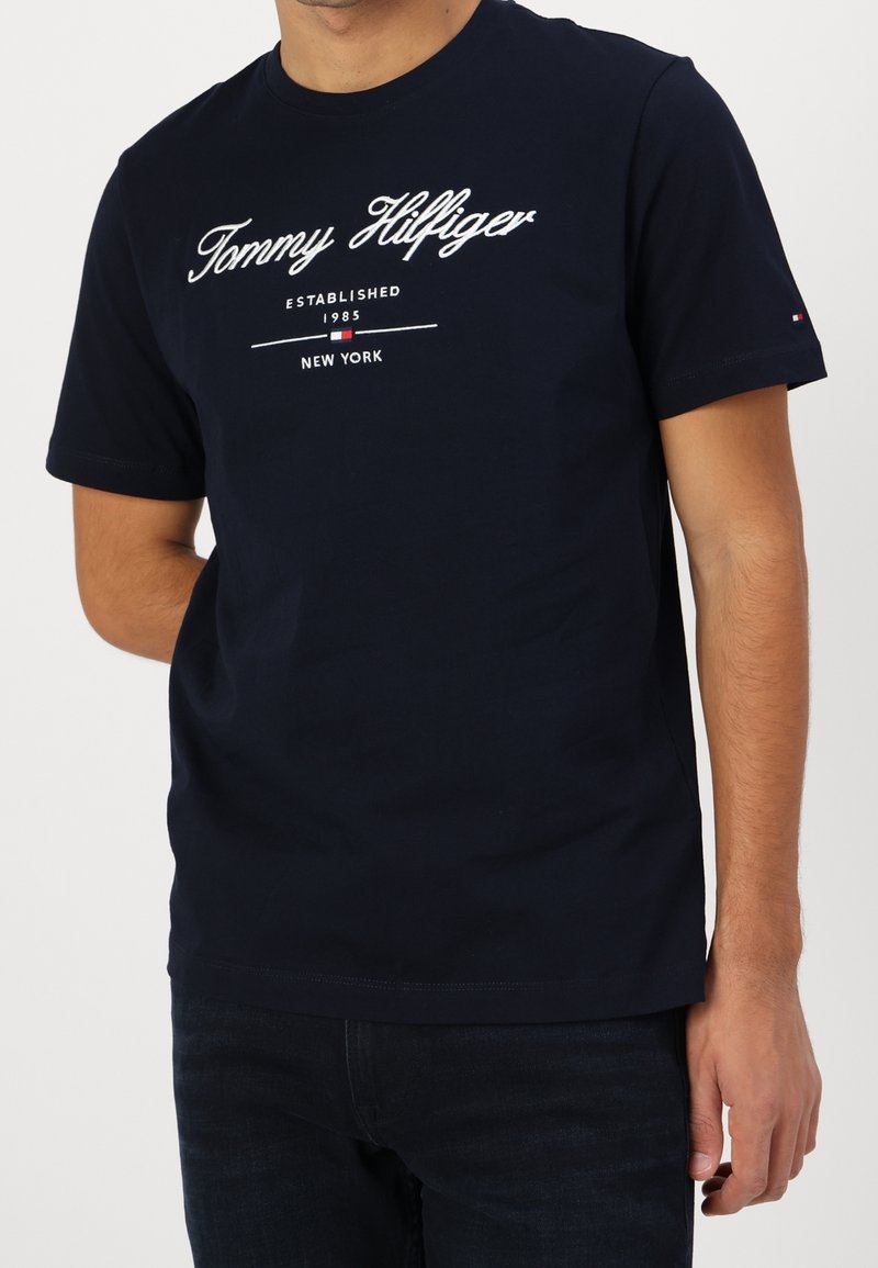 Homme portant un t-shirt Tommy Hilfiger bleu marine avec un texte de logo blanc et un petit emblème de drapeau sur la manche.