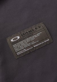 Čierna textilná etiketa s textúrovaným povrchom, zobrazujúca názov značky "Oakley" a výrobné detaily bielym a strieborným textom. Obdĺžnikový tvar.