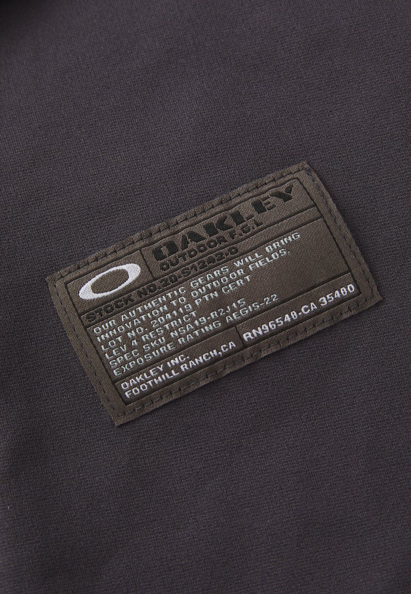 Etichetta in tessuto nero con superficie testurizzata, che mostra il nome del marchio "Oakley" e i dettagli di produzione in testo bianco e argento. Forma rettangolare.