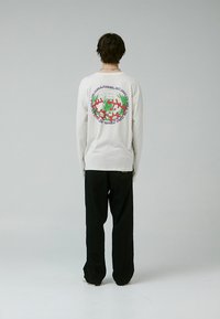 Camiseta blanca de manga larga con un gráfico colorido en la espalda, combinada con pantalones negros y una etiqueta visible en la cinturilla.