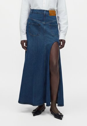 Femme portant une longue jupe en jean bleu avec une fente devant et des poches plaquées, associée à des ballerines noires pointues et une chemise blanche à manches longues.