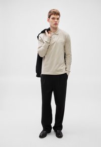 Bež dolg rokav polo majica s trirazširno zaponko, kombinirana s črnimi hlačami. Model ima črno jakno preloženo čez ramo.