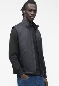 Schwarze ärmellose Jacke mit hohem Kragen, Reißverschluss, Seitentaschen und glatter Textur, getragen über einem schwarzen Langarmshirt.