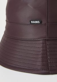 Ljubičasta vodootporna kapa s širokim rubom, glatke teksture, sa šivanim detaljima i malom crnom oznakom RAINS.