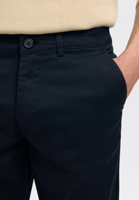 Nahaufnahme von dunkelblauen Hosen mit Knopf und Gürtelschlaufe, Hand teilweise in der Vorderhose, beiger strukturierter Oberteil an der Taille sichtbar.