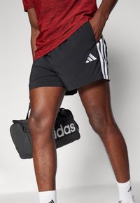 Shorts de sport noirs avec des rayures blanches, un t-shirt rouge texturé et des chaussettes blanches. Un sac de sport noir avec le logo "adidas" est également visible.