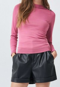 Kobieta w dopasowanej różowej bluzce z długim rękawem i detalami w postaci guzików oraz czarnych szortach z wysokim stanem z ekoskóry, trzymająca ręce w kieszeniach.
