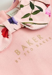 Top de algodón rosa con un collar floral con patrones en púrpura, rojo y verde. El nombre de la marca está grabado en dorado en el centro.