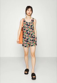 Macacão sem mangas com padrão floral, apresentando folhagem verde e flores cor-de-rosa sobre um fundo azul-marinho; inclui bolsos laterais. Sandálias plataforma pretas e uma bolsa laranja.