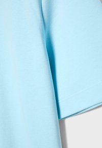 Zbliżenie na jasnoniebieską bawełnianą tkaninę z widocznymi szwami i przeszyciami, pokazujące rękaw i część ciała krótkiego rękawa ubrania.